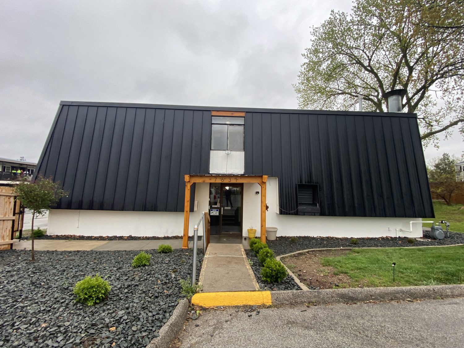 A cedar framed entrance at Urban Heights, set into the black metal facade above a white painted base, with a concrete walk, river rock beds and new shrubs on either side.