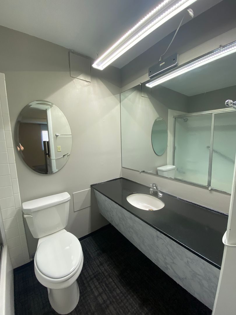 A bathroom with a dark stone countertop, a round mirror, a light bar above the sink and a glass shelf.