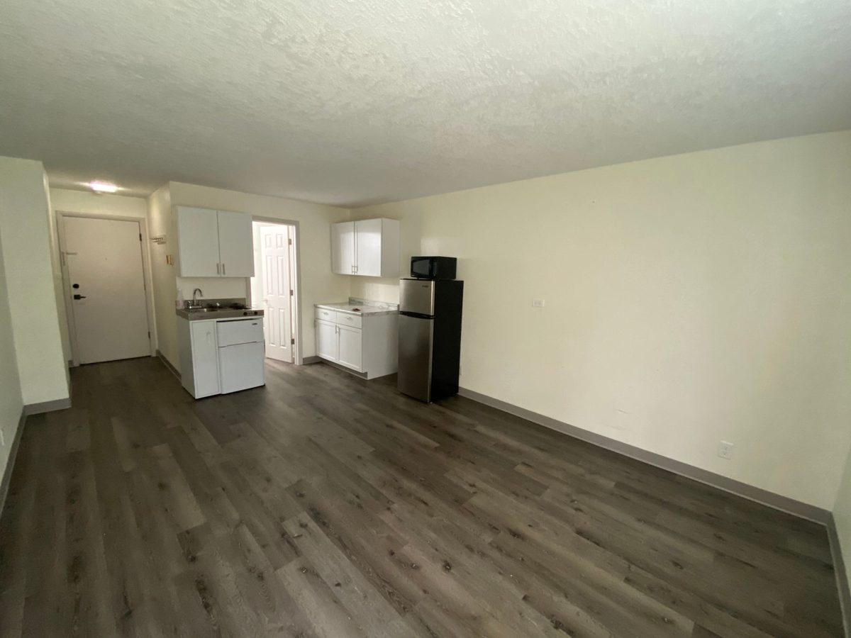 An empty studio with plank flooring, a white kitchenette with upper cabinets, and the entry door to the right.