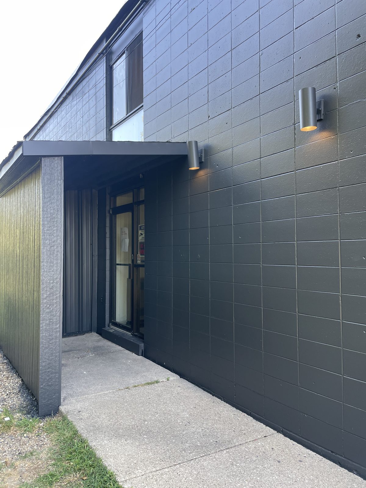 The corner of the building, clad in dark charcoal panels, with an entry canopy and exterior downlights above the door.