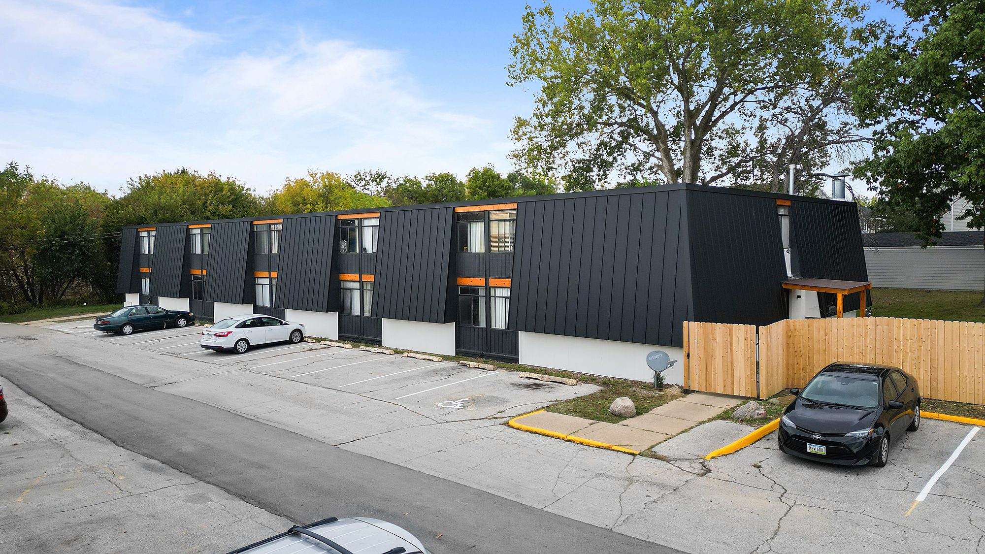 Urban Heights from above, a long two story apartment building clad in black standing seam metal over a white painted base, with cedar framed entries and a striped parking lot running the length of the building.